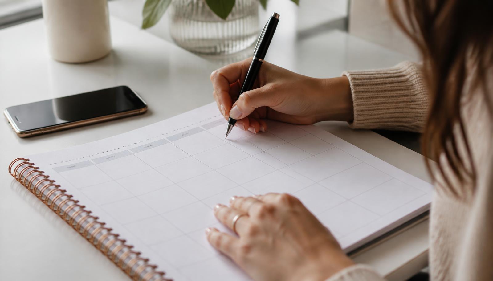 Hands writing a recurring wellness appointment into a weekly planner beside a phone